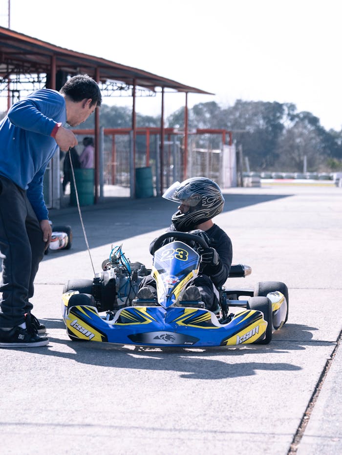 A child in a go-kart being guided by an adult at an outdoor racing track, wearing a helmet.